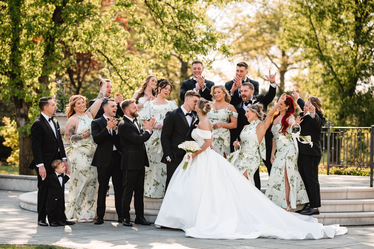 Lively wedding party celebrating outdoors with bridesmaids in floral dresses and groomsmen in black tuxedos.