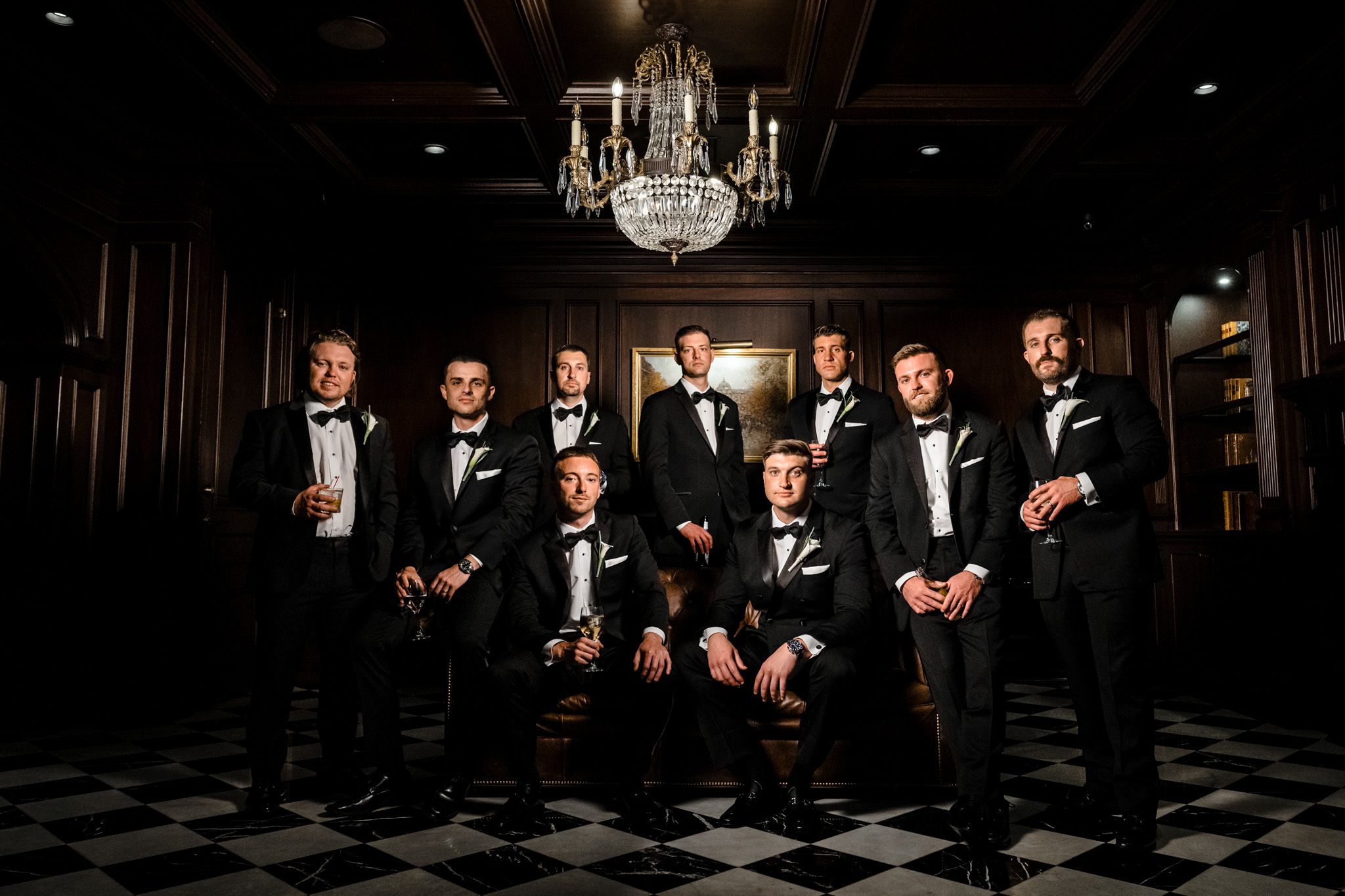 Groomsmen in black tuxedos posing under a crystal chandelier on a checkered floor.