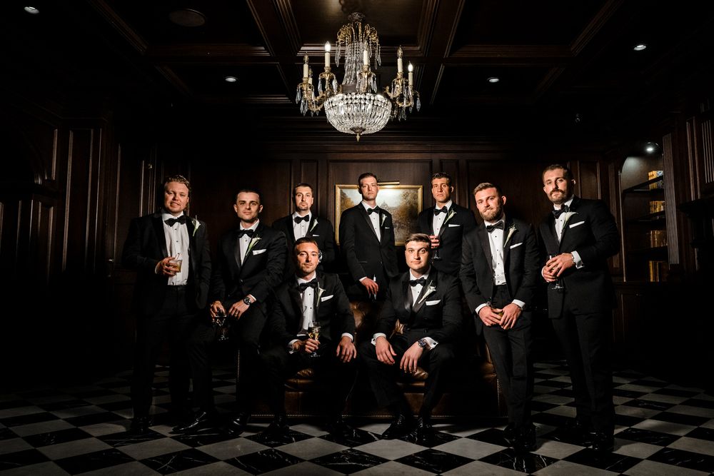 Groomsmen in black tuxedos posing under a crystal chandelier on a checkered floor.