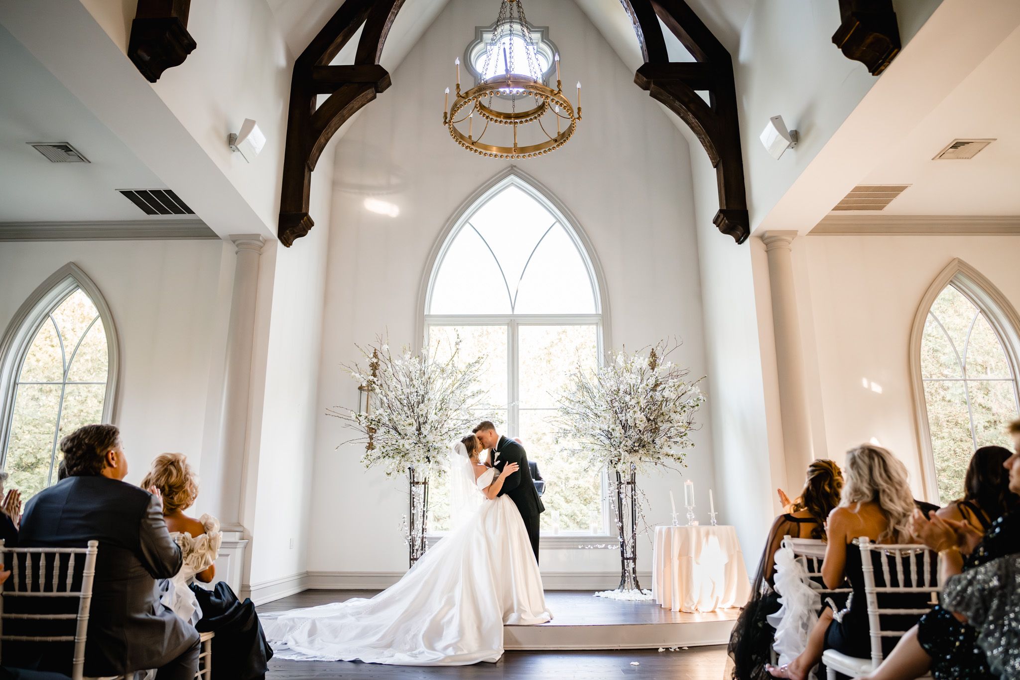 Bride and groom sharing first kiss at altar in stunning white chapel with gothic arched windows.