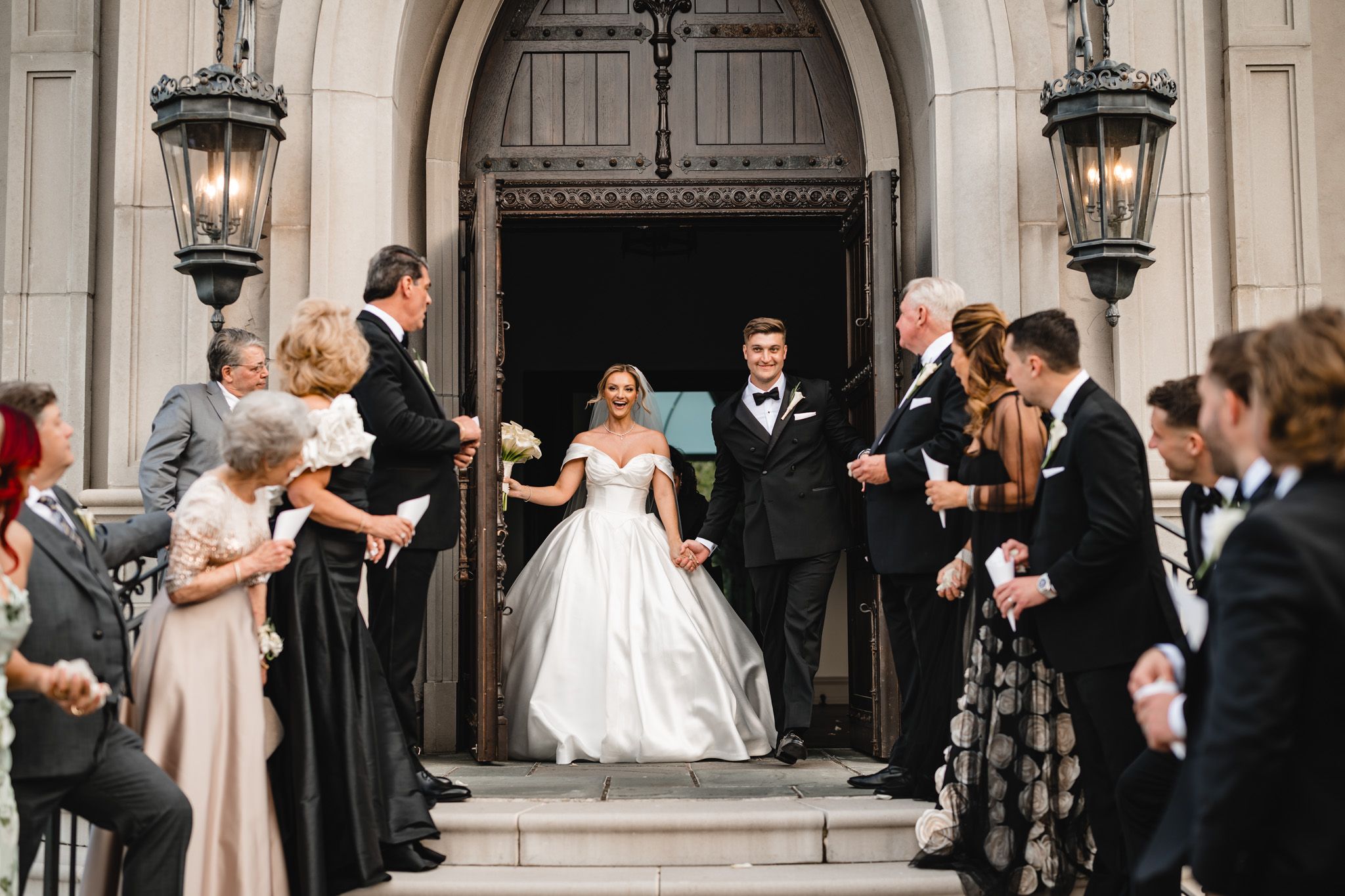 Smiling bride and groom exit historic church doors surrounded by celebrating wedding guests.