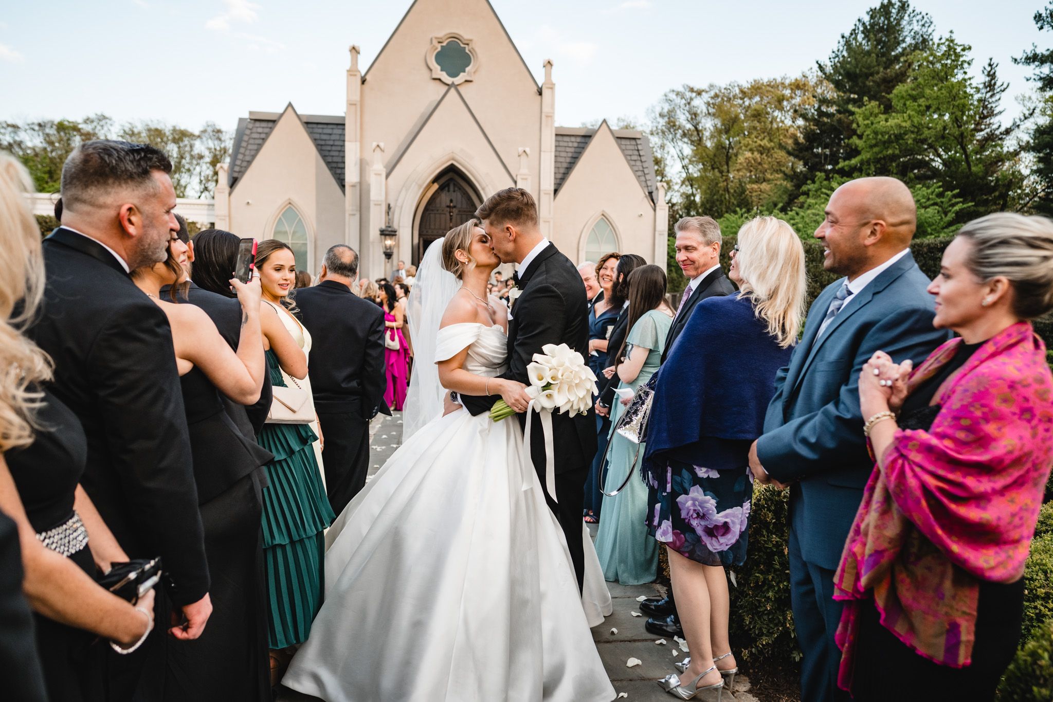 Newlyweds share a first kiss outside a stone chapel surrounded by wedding guests in formal attire.