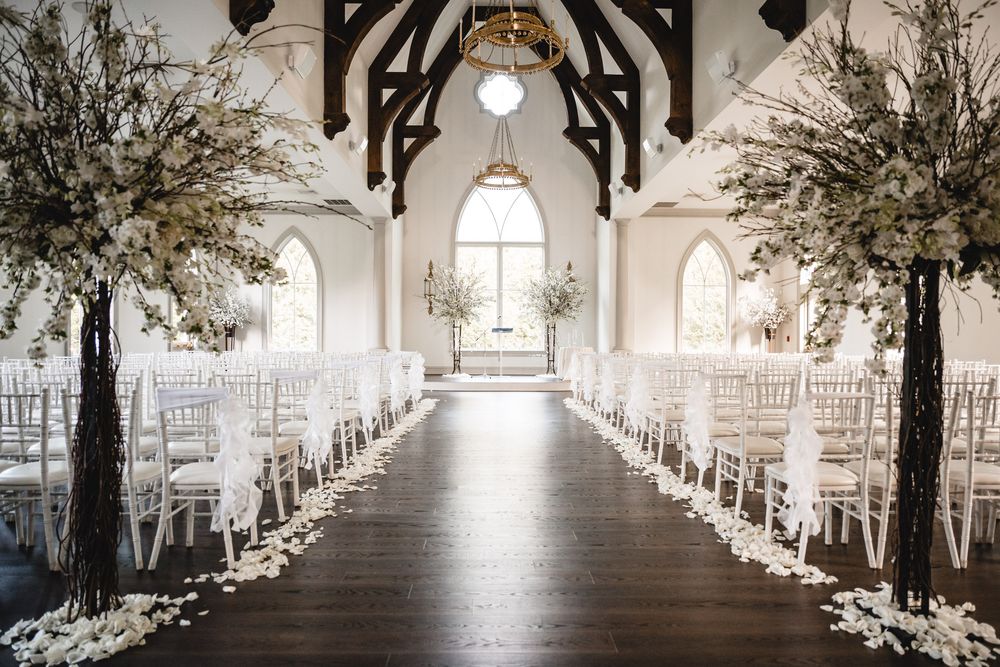 Elegant wedding ceremony aisle with white chairs and tall floral arrangements in a chapel.