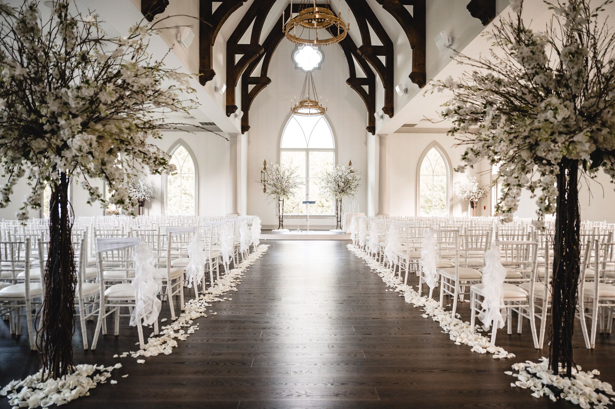 Elegant wedding ceremony aisle with white chairs and tall floral arrangements in a chapel.