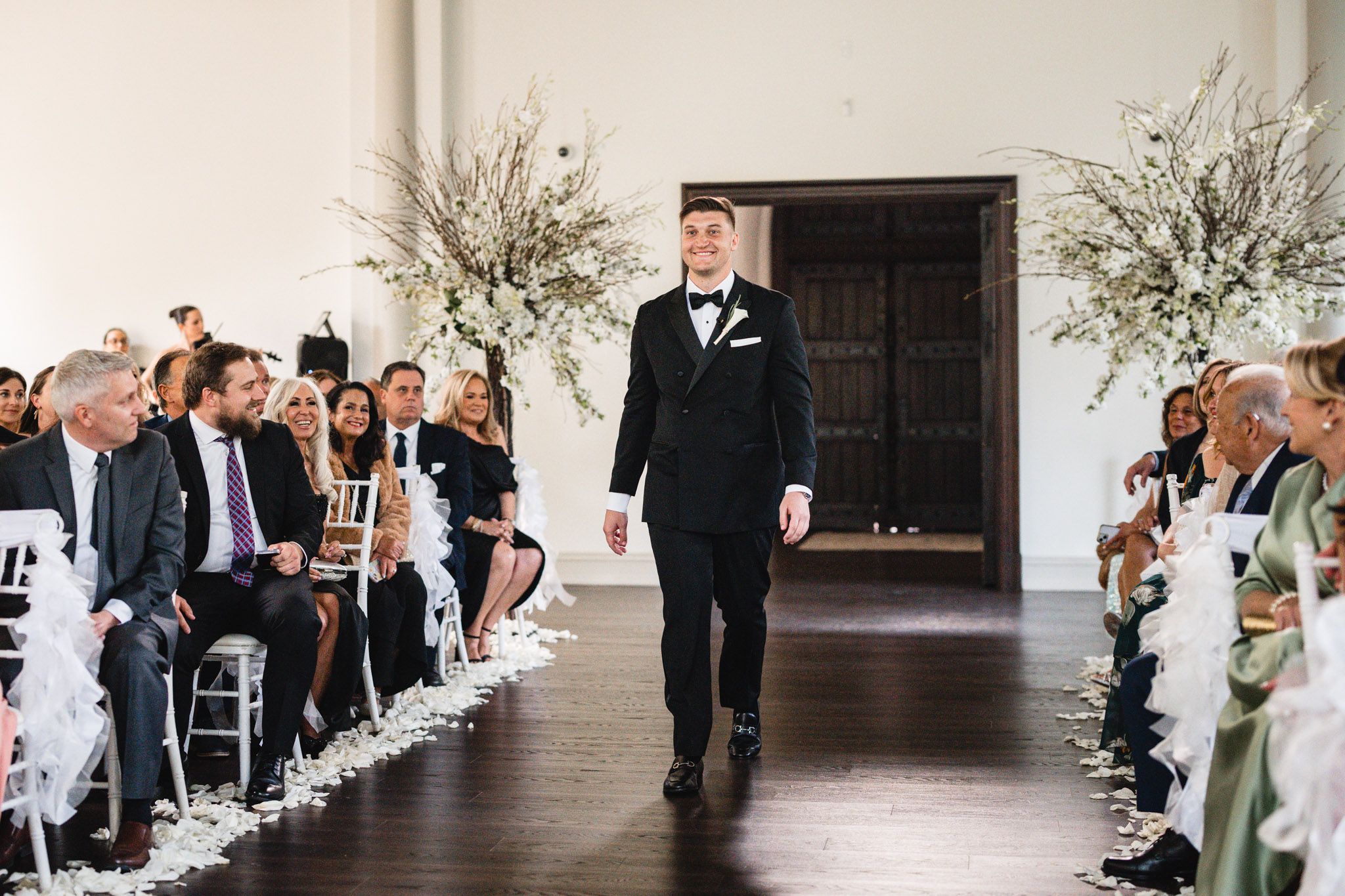 Groom in black tuxedo walking down the aisle of a bright white wedding ceremony venue.