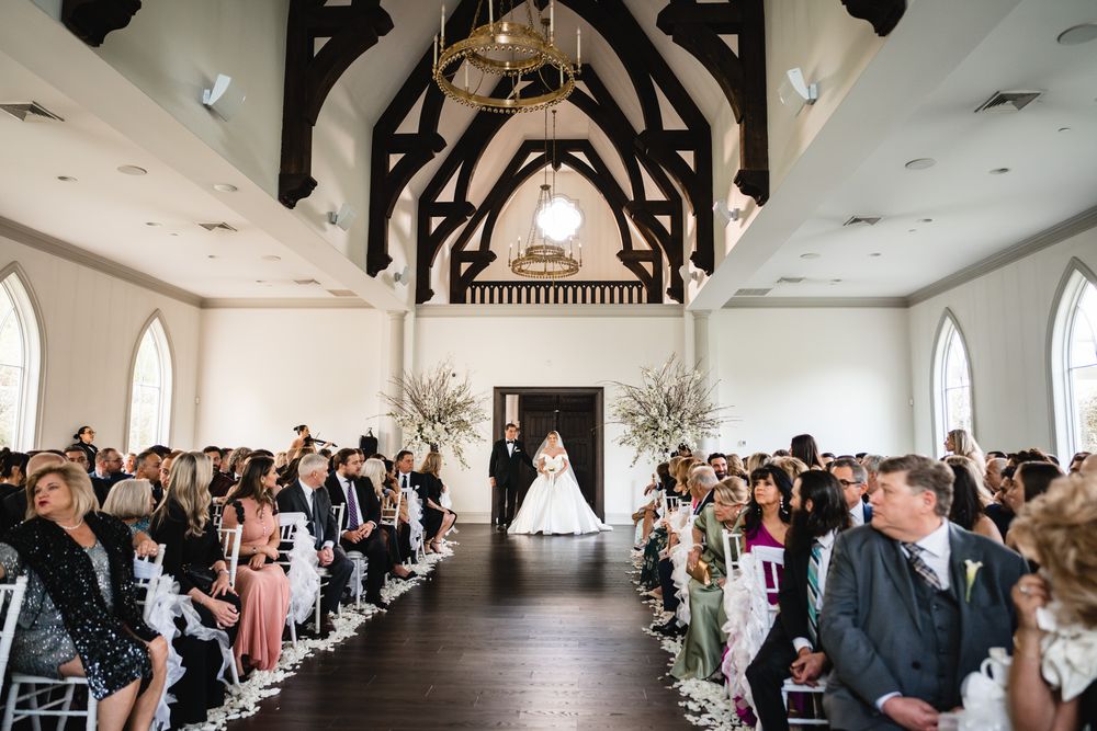 Wedding ceremony inside a white chapel with vaulted ceilings, floral arrangements, and seated guests.