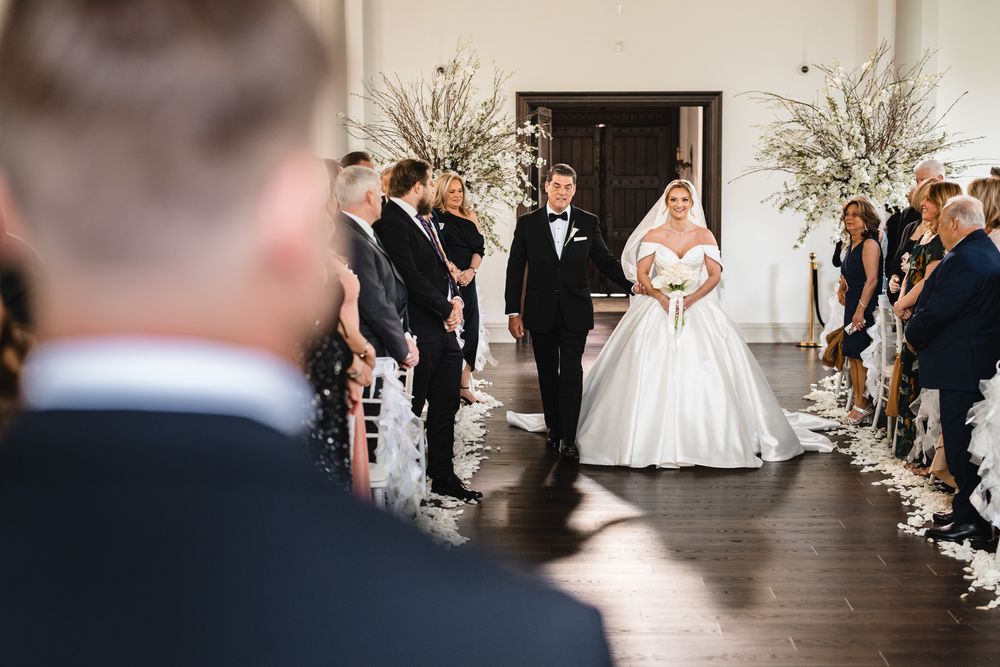 Bride in voluminous white ball gown walking down aisle with escort during elegant wedding ceremony.