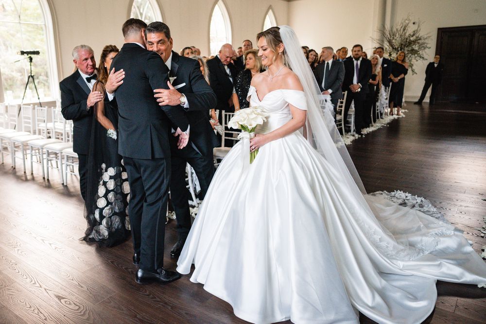 A bride in a stunning white ball gown with veil greets guests during an elegant chapel ceremony.