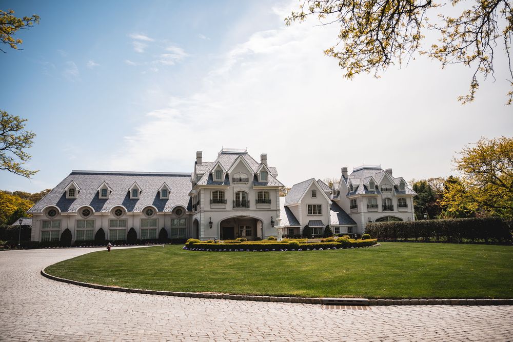 Grand white French chateau-style mansion with manicured lawn and circular cobblestone driveway.