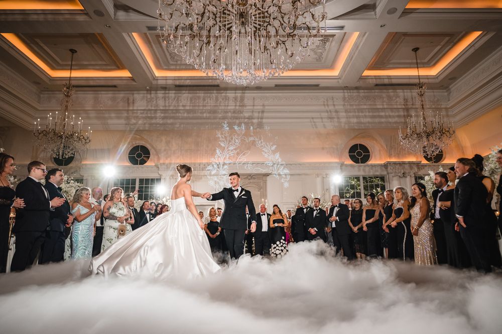 Bride and groom share first dance surrounded by fog and guests in elegant ballroom with chandeliers.
