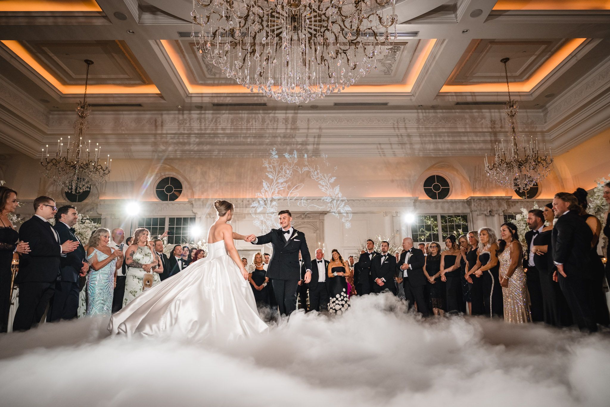 Bride and groom share first dance surrounded by fog and guests in elegant ballroom with chandeliers.