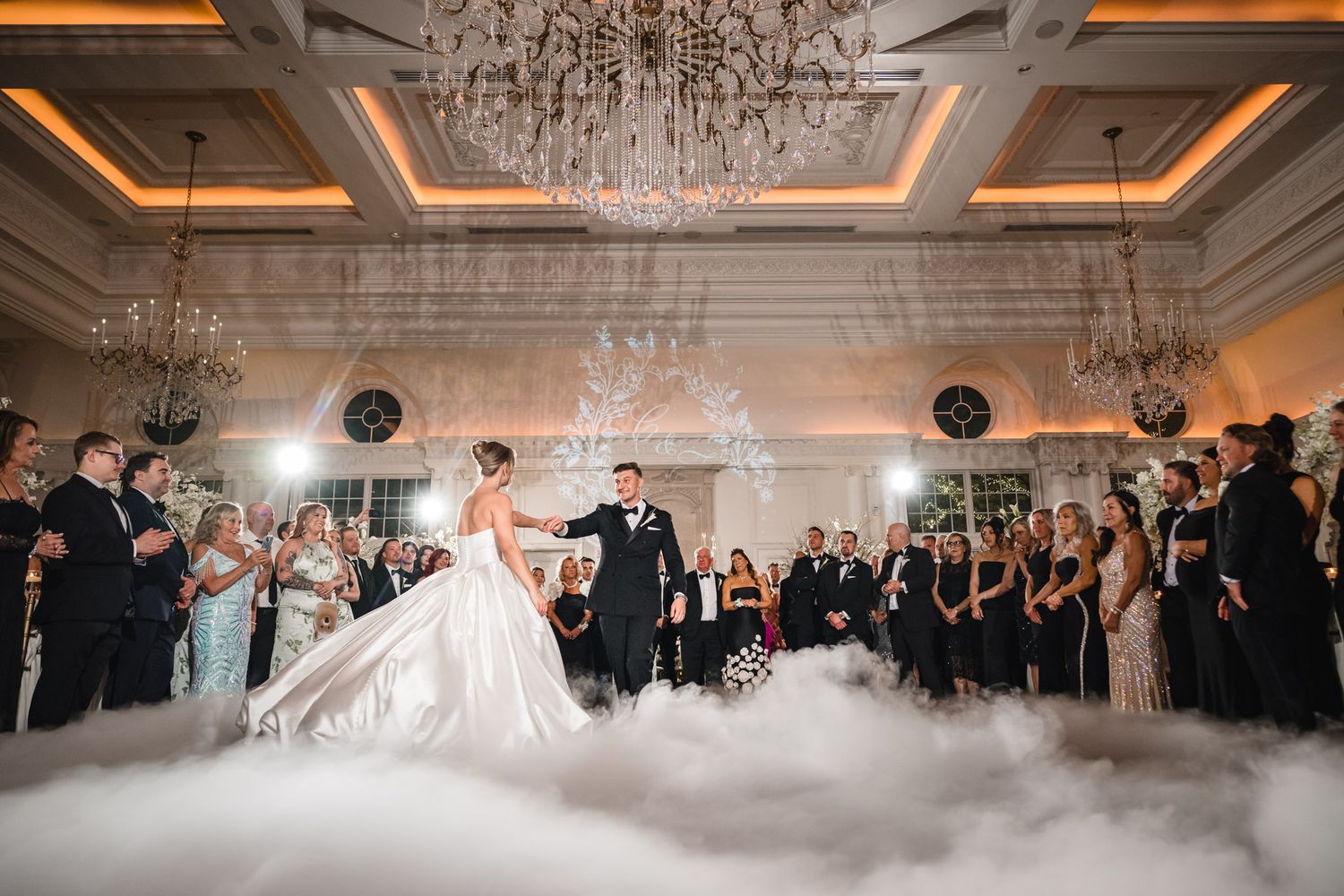 Bride and groom share first dance surrounded by fog and guests in elegant ballroom with chandeliers.