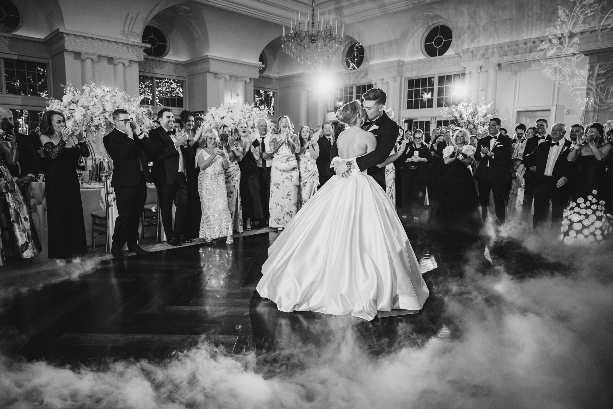 Bride and groom share first dance surrounded by guests in a dreamy fog-filled ballroom.
