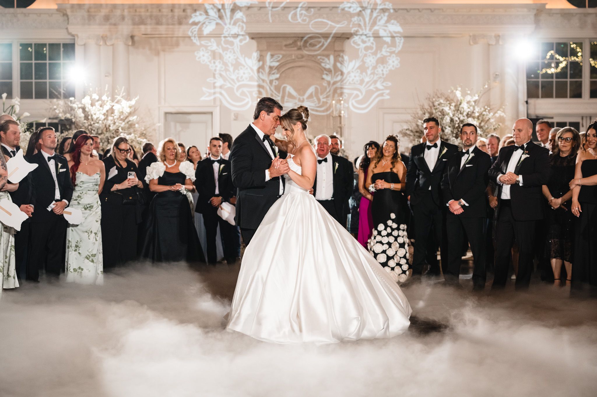 Bride and groom share first dance surrounded by guests in elegant ballroom with dreamy fog effect.