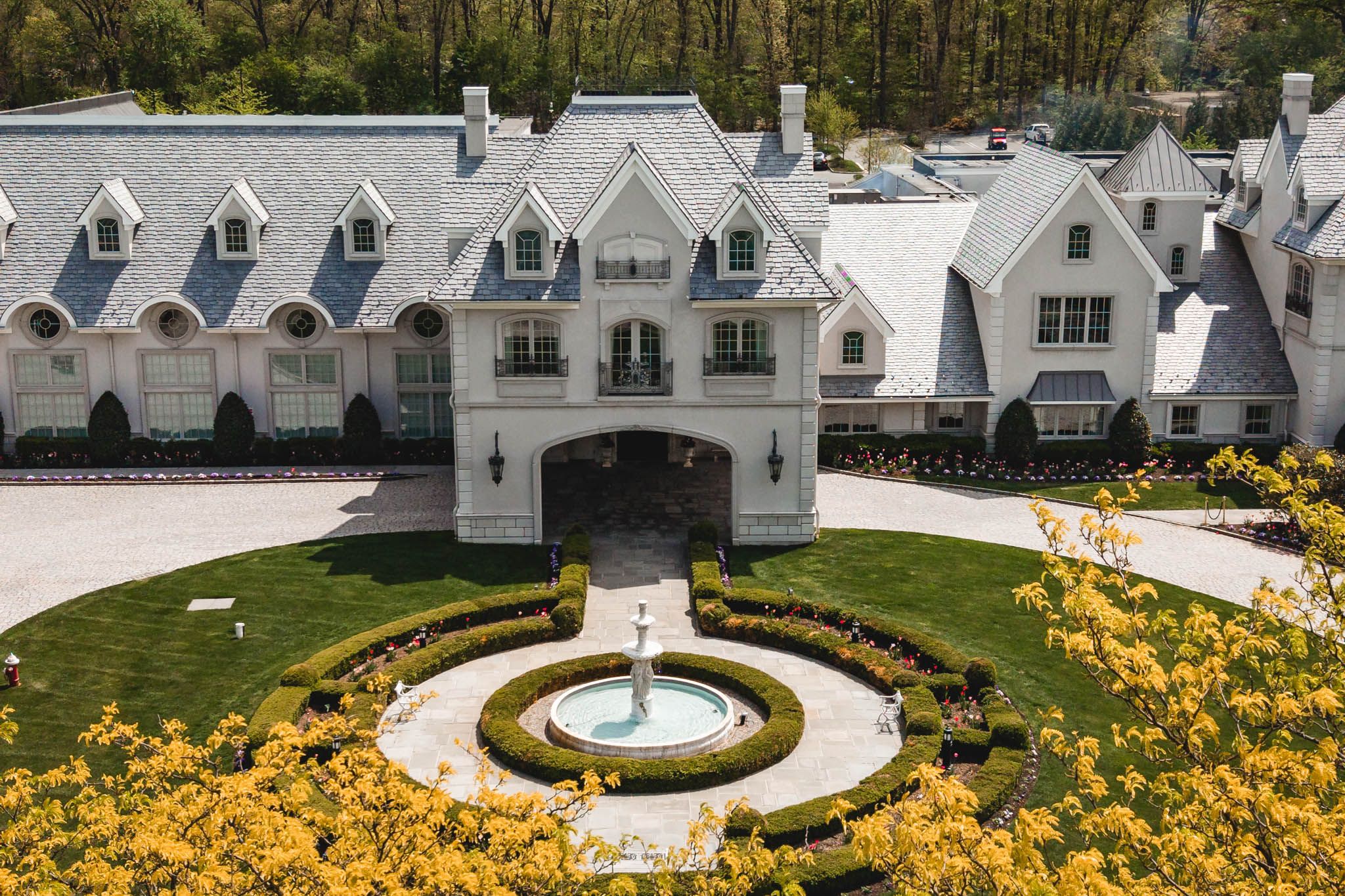 Aerial view of a grand estate mansion with fountain, manicured lawn, and yellow flowering shrubs.