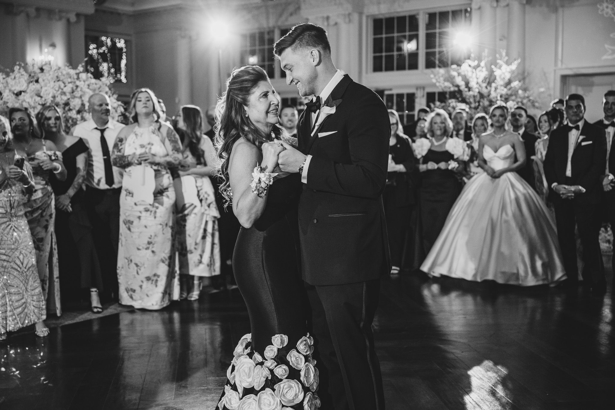 Bride and groom share first dance at elegant wedding reception while guests watch in black and white photo.