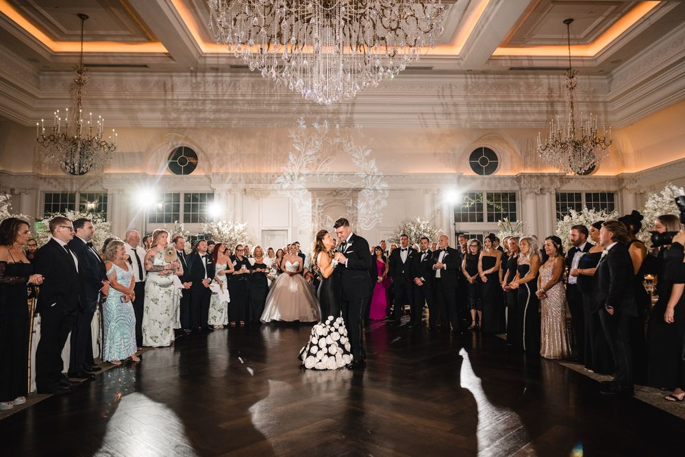 Couple shares romantic first dance under crystal chandeliers surrounded by wedding guests in grand ballroom.