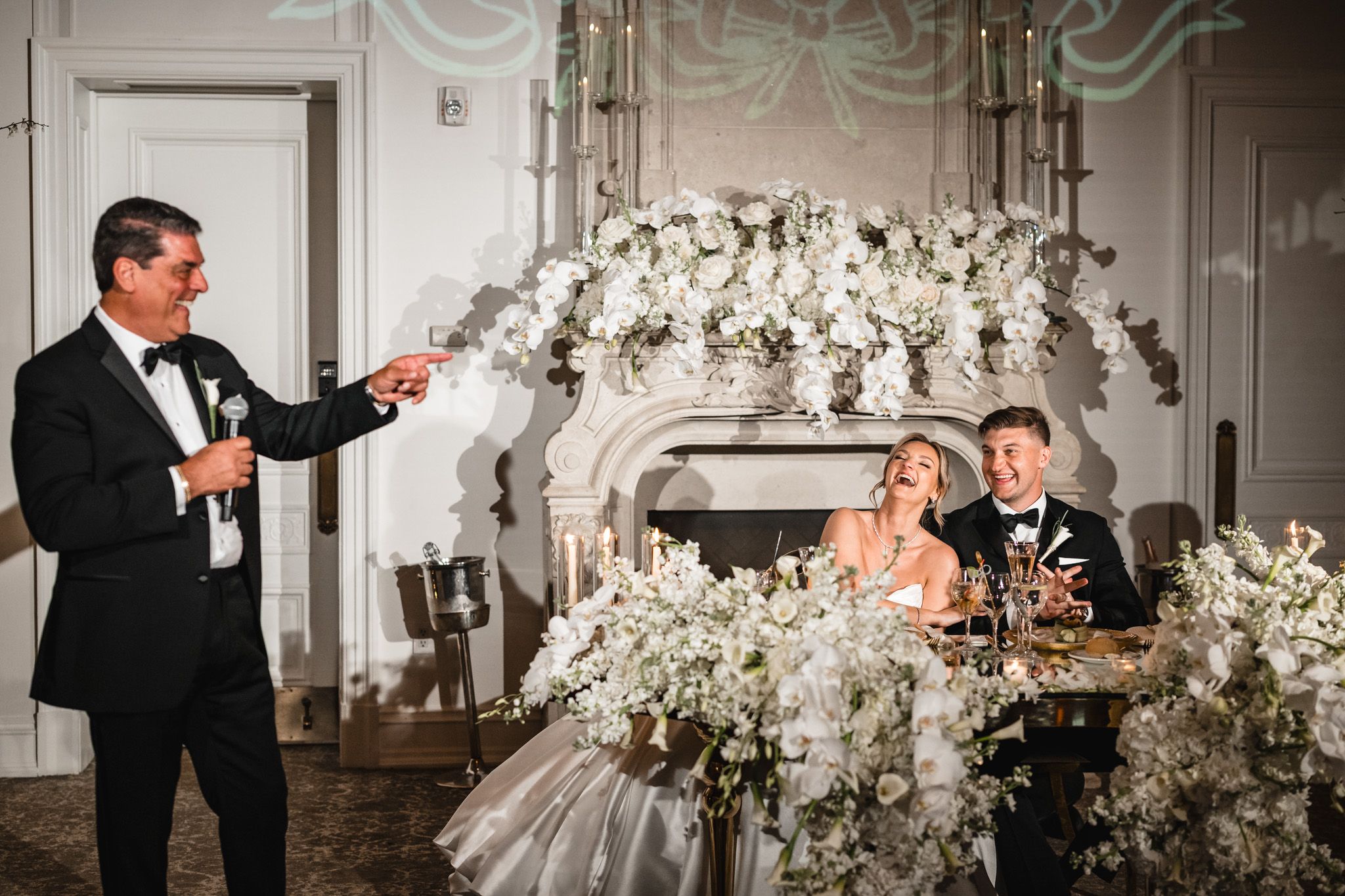 Man giving wedding toast beside bride and groom at elegant white floral reception table near ornate fireplace.