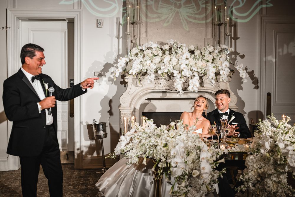 Man giving wedding toast beside bride and groom at elegant white floral reception table near ornate fireplace.