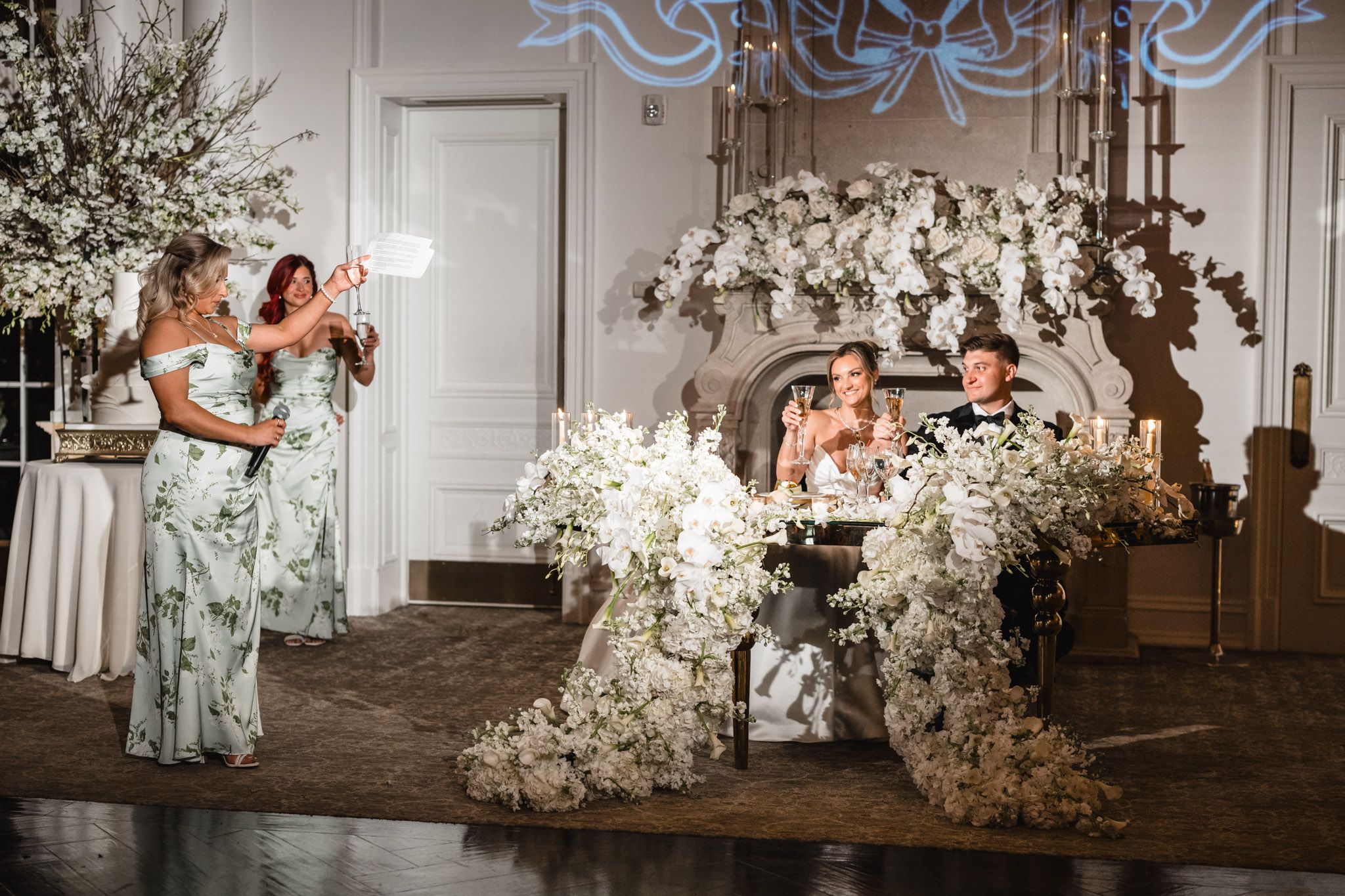 Bridesmaid giving a toast at a wedding reception with white floral arrangements and sweetheart table.