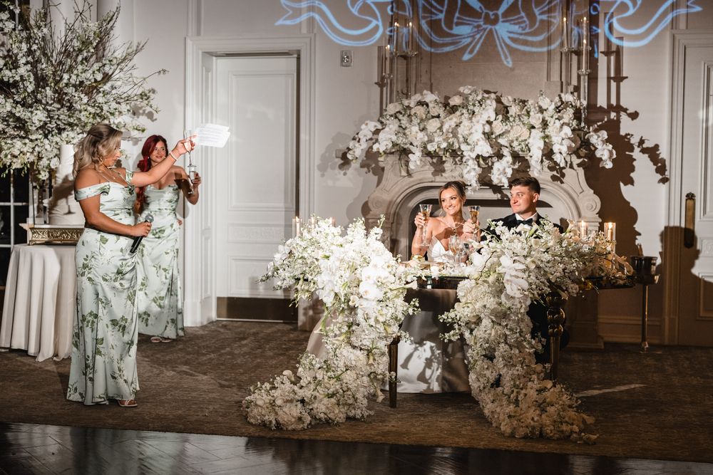 Bridesmaid giving a toast at a wedding reception with white floral arrangements and sweetheart table.