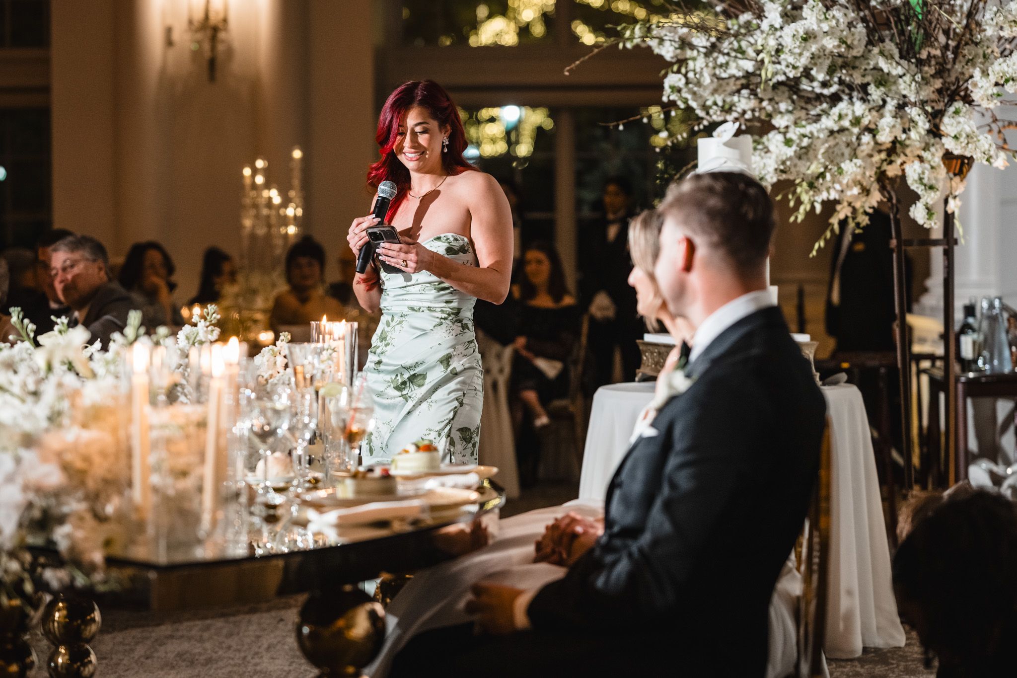 Woman with red hair gives speech at elegant wedding reception with floral arrangements and candlelight.