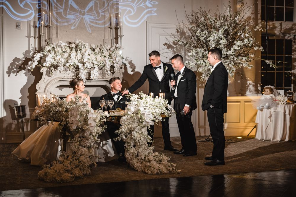 Groomsmen gather around the sweetheart table adorned with white floral arrangements at a wedding.