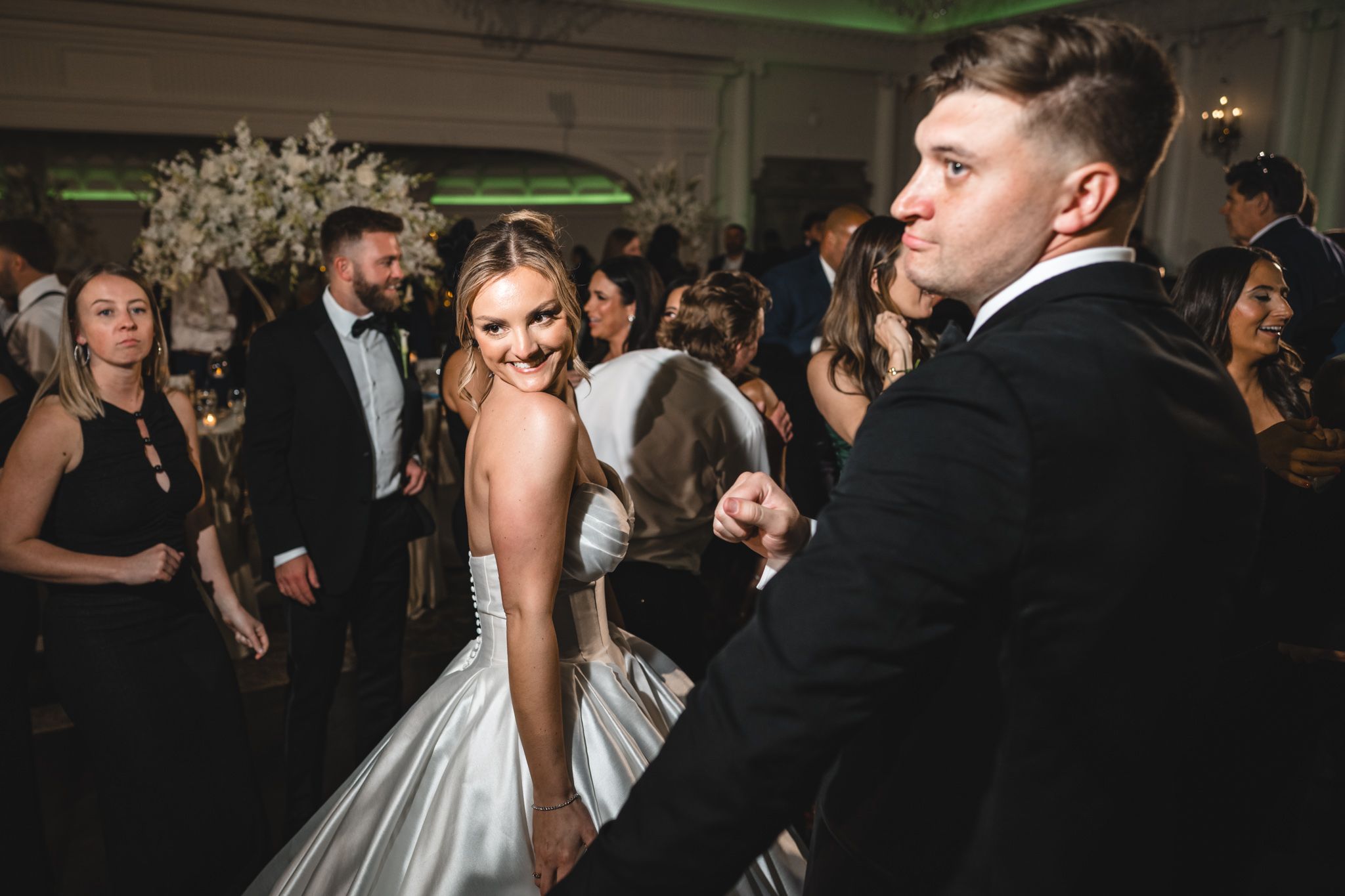 Bride in silver gown smiles at groom in tuxedo while dancing at elegant wedding reception.
