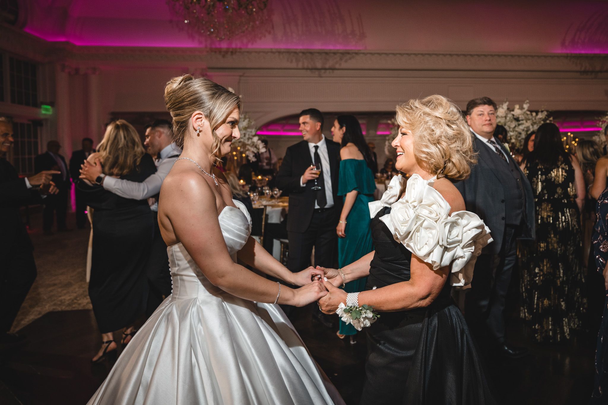 Bride in white satin gown dances joyfully with older woman in black dress at wedding reception.
