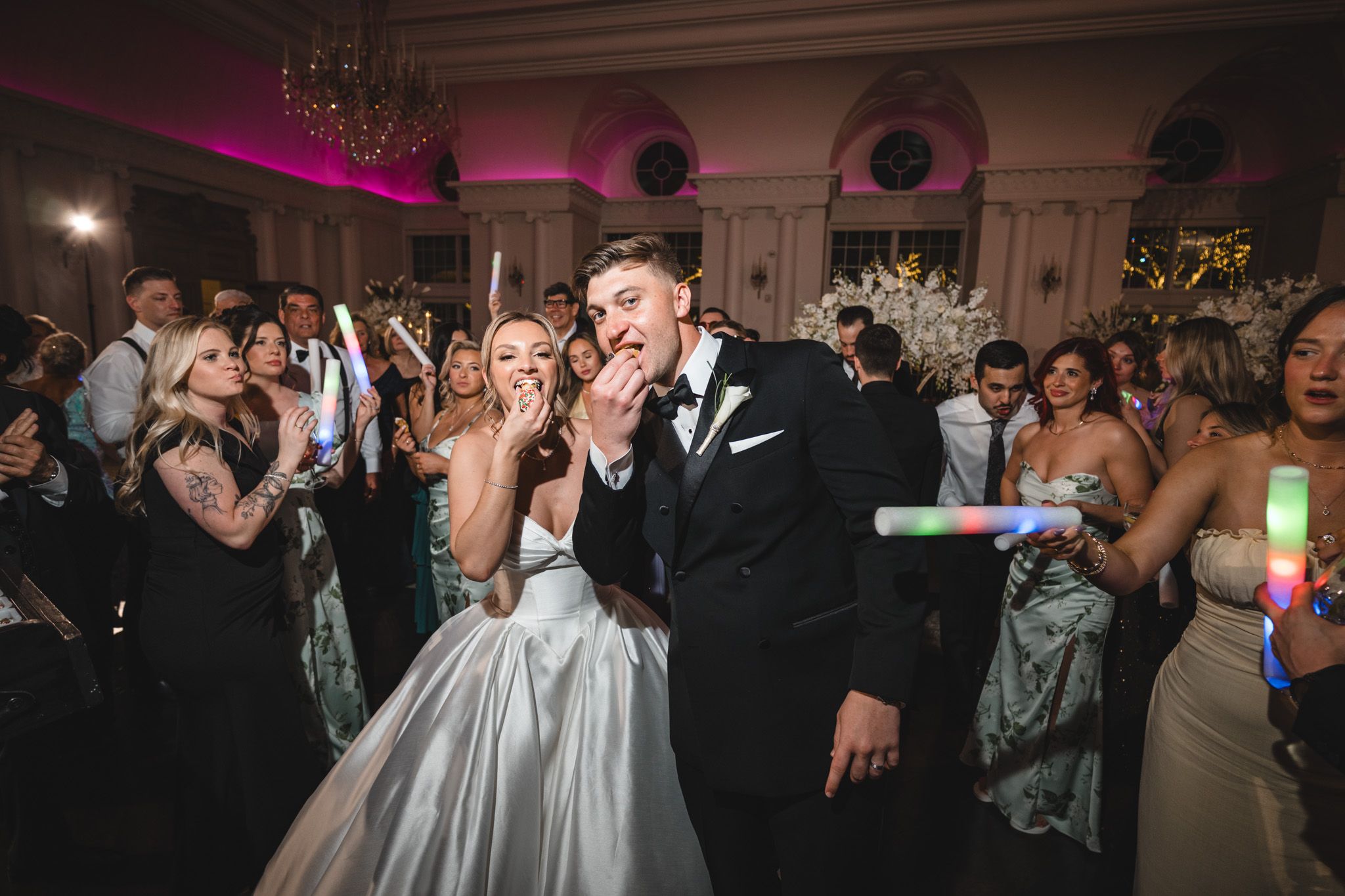 Excited bride and groom celebrate with glowing sticks on dance floor surrounded by cheering wedding guests.