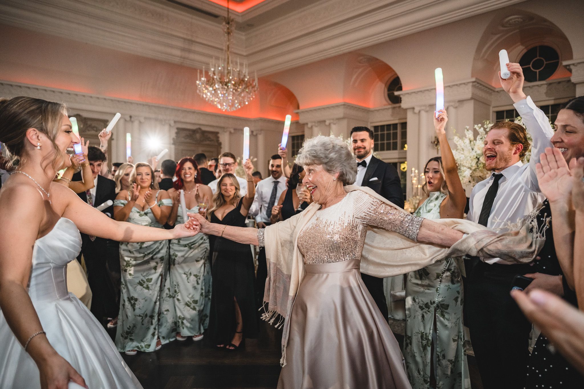 Elderly woman dances joyfully with guests holding light sticks at a grand wedding reception.