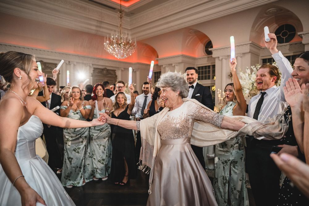 Elderly woman dances joyfully with guests holding light sticks at a grand wedding reception.