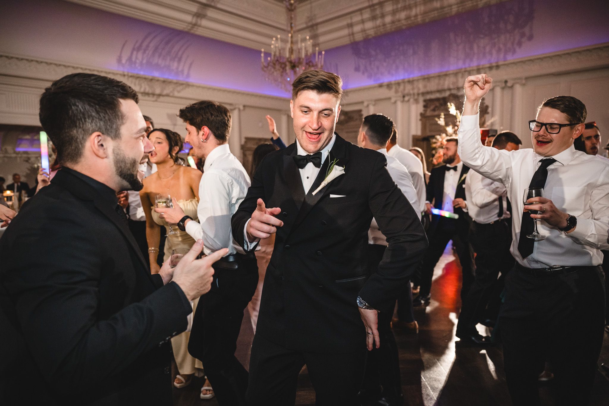 Groom in black tuxedo celebrates on dance floor with guests at wedding.