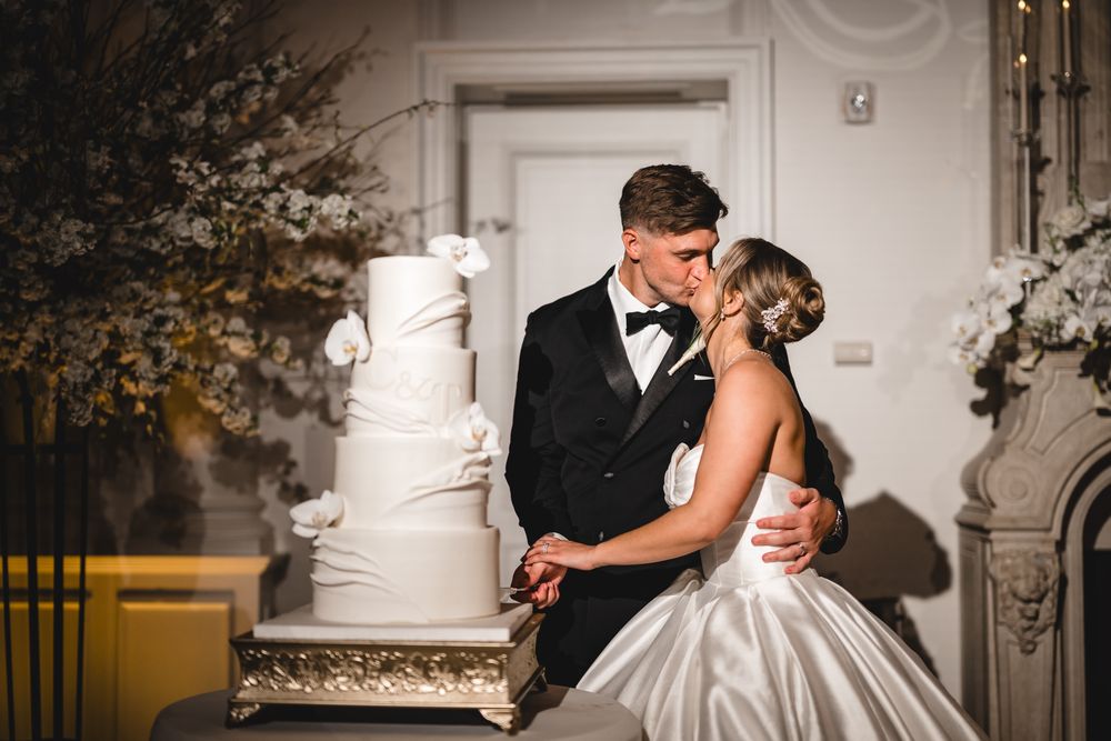 Bride and groom share romantic kiss beside elegant white tiered wedding cake.