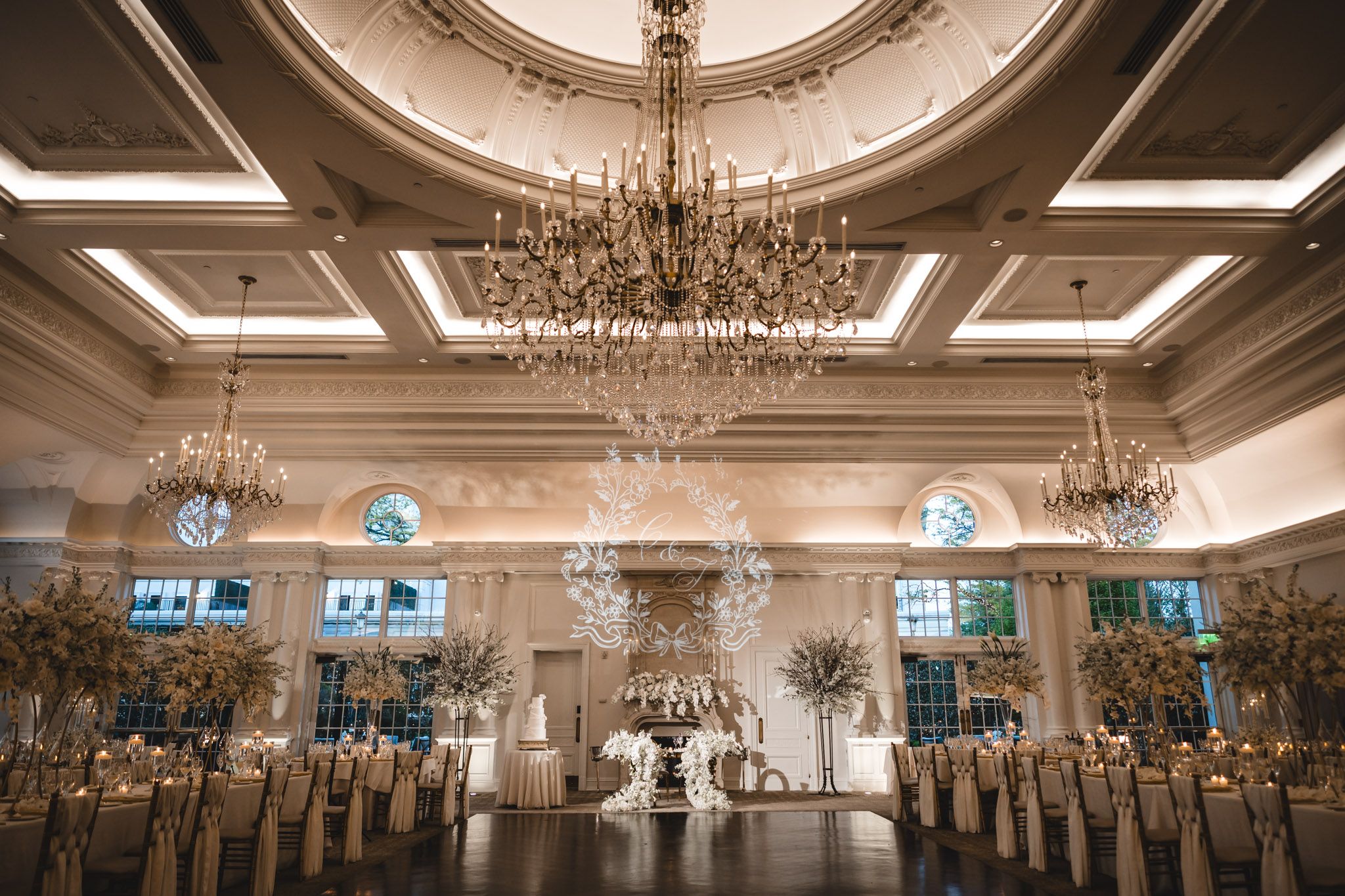 An elegant wedding reception ballroom features grand crystal chandeliers and tall floral centerpieces.