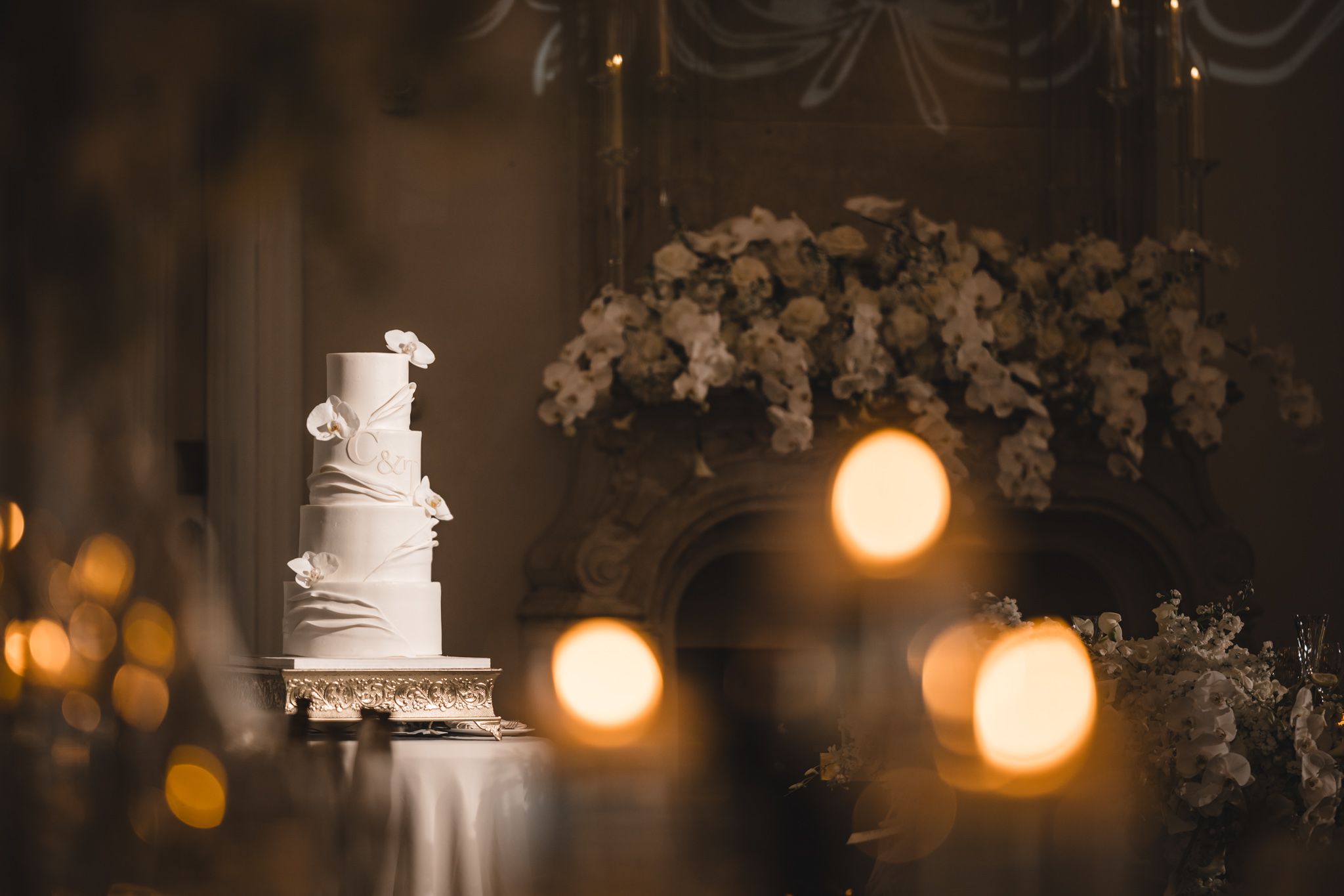 A four-tier white wedding cake with floral details glows beside candles and flower arrangements.