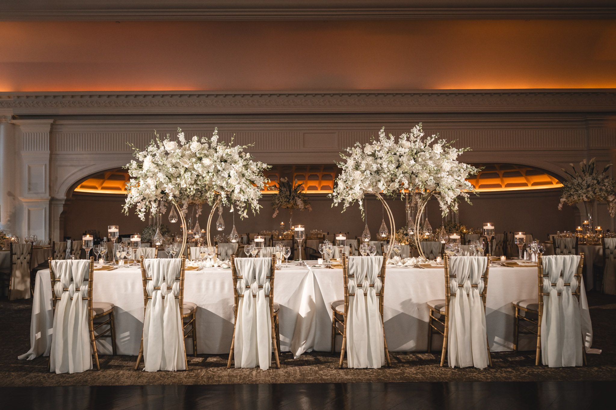 A sweetheart table with tall white floral tree centerpieces and draped chairs in a grand ballroom.