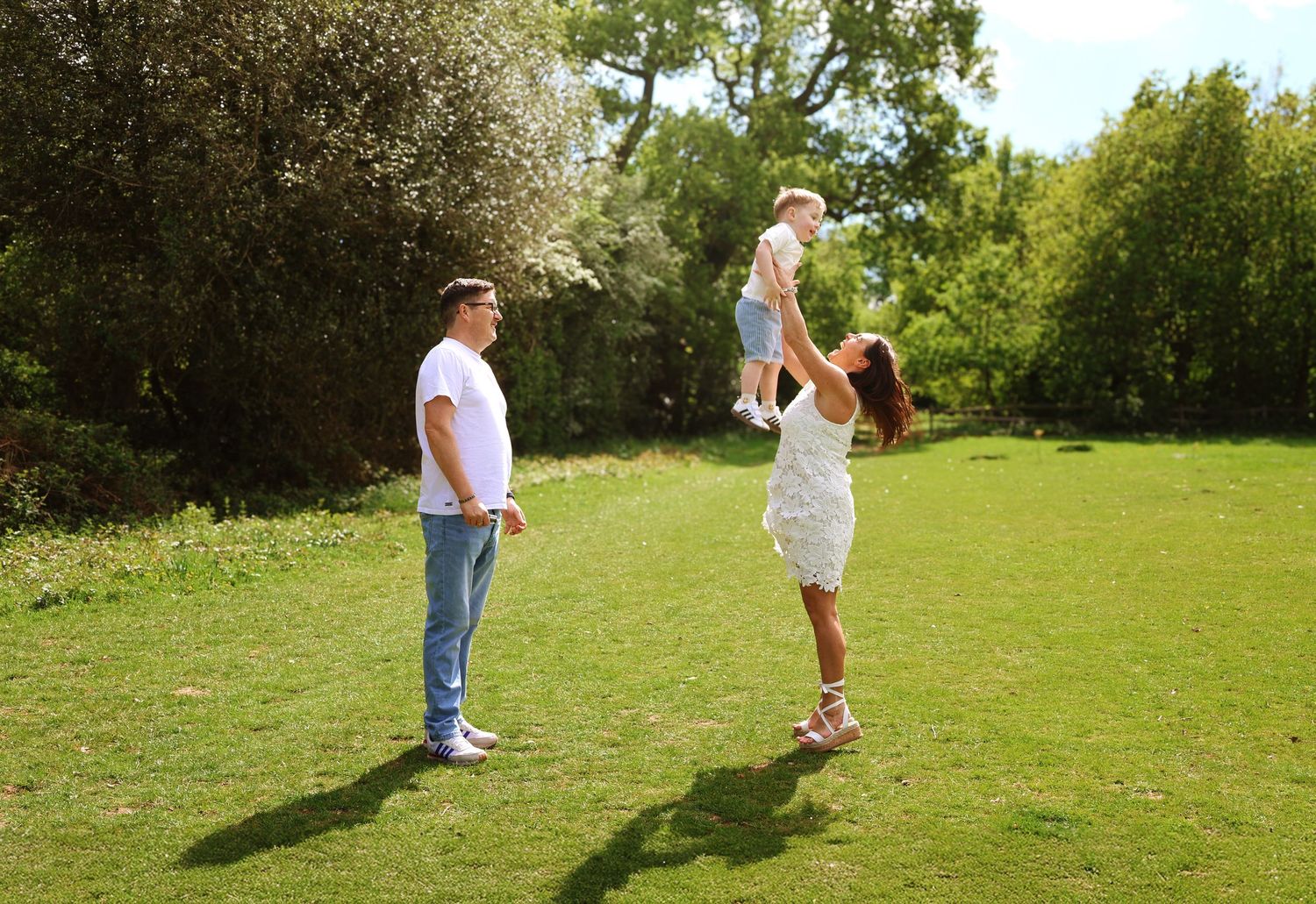 A happy family outdoors, with a woman lifting a baby in the air while a man watches on a sunny green lawn.