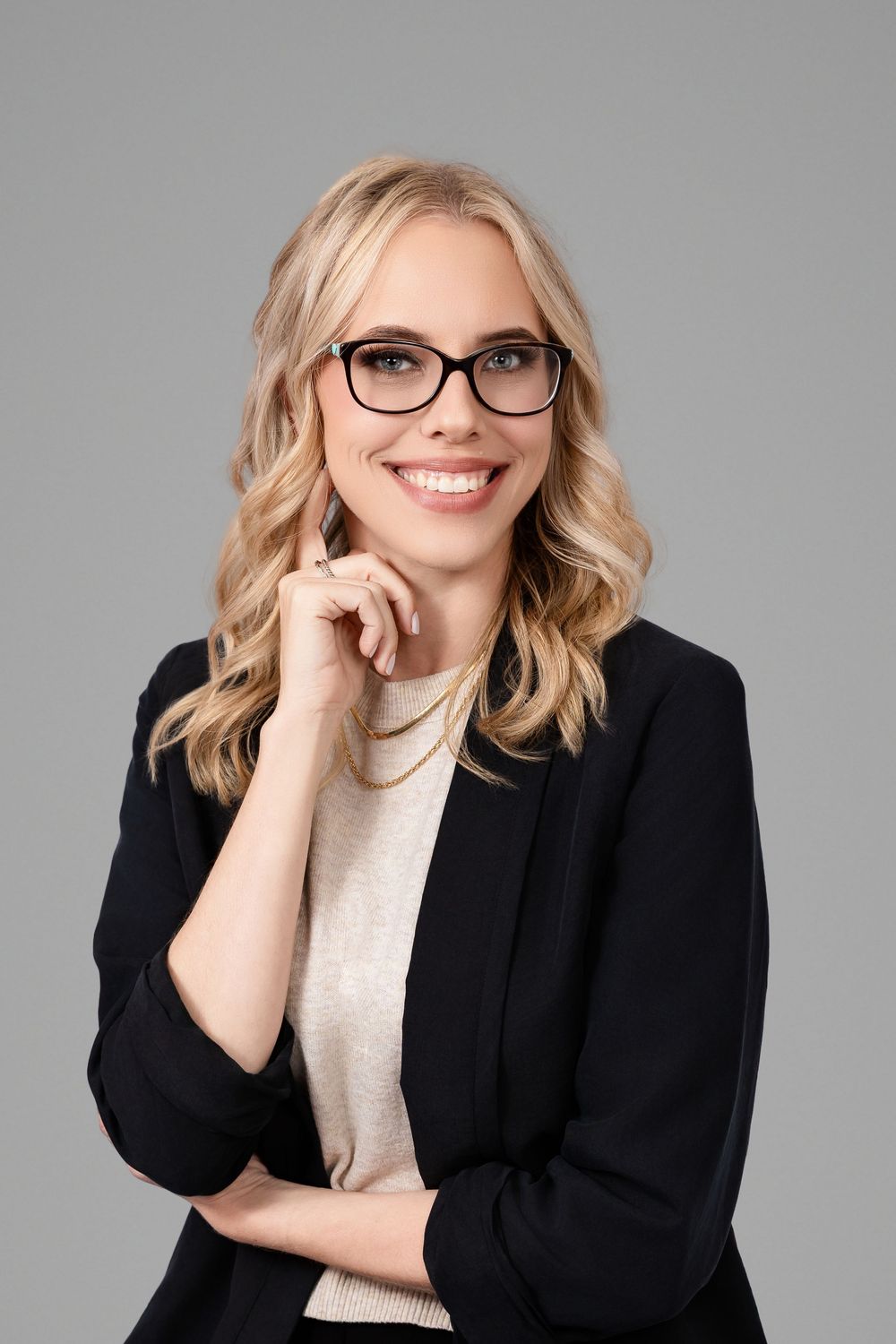 Smiling blonde woman in black blazer and glasses posing confidently against a gray background.