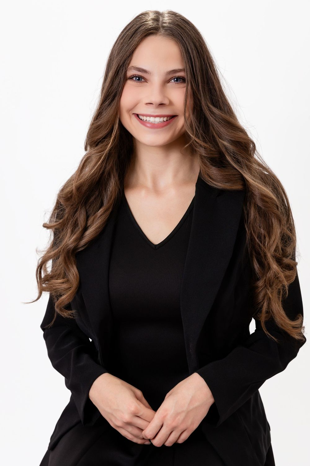 Smiling young woman with long wavy brown hair wearing a black blazer and top, posing against a white background.