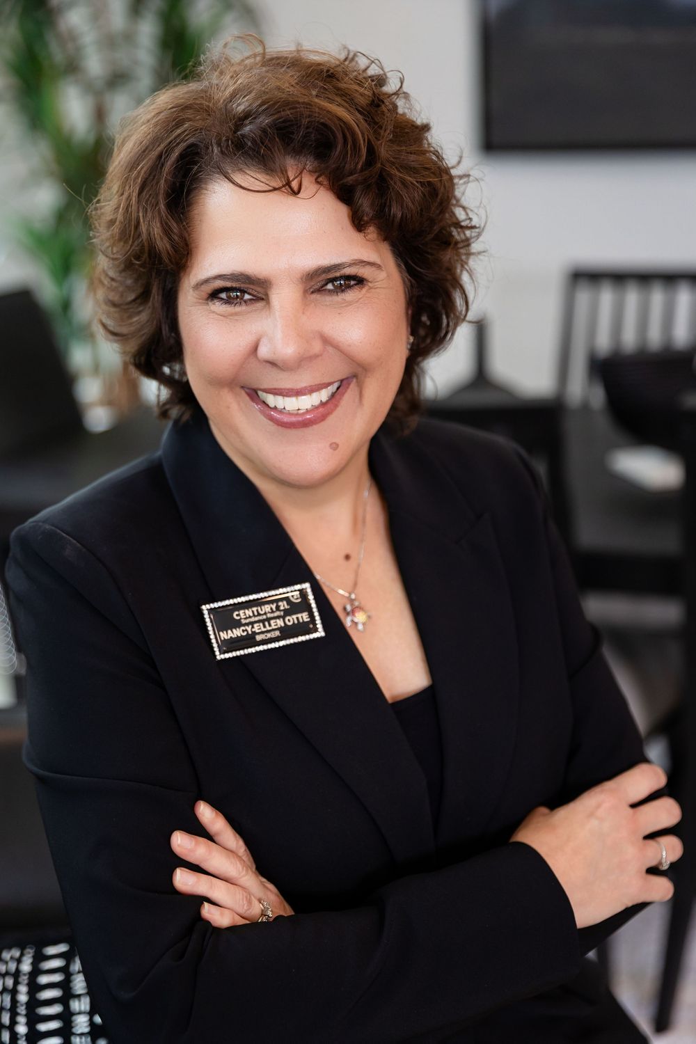 Smiling Century 21 real estate agent Nancy Ellen Otis in black blazer with name badge, arms crossed, professional headshot.