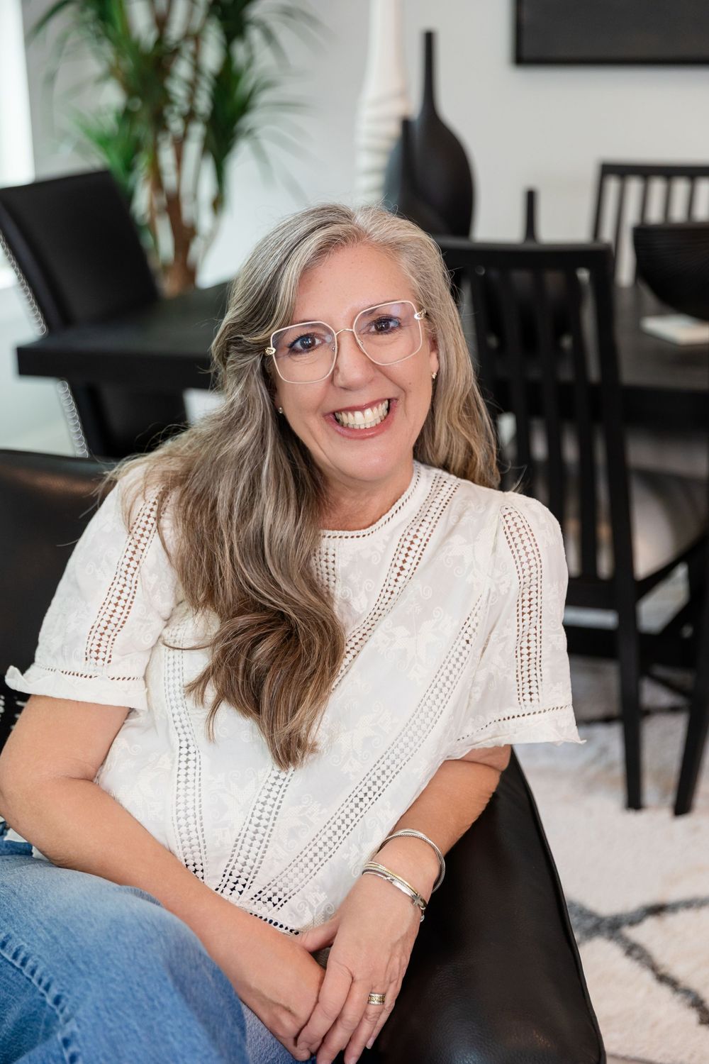 Smiling middle-aged woman with long gray hair and pink glasses wearing a white lace top, seated in a modern living room.