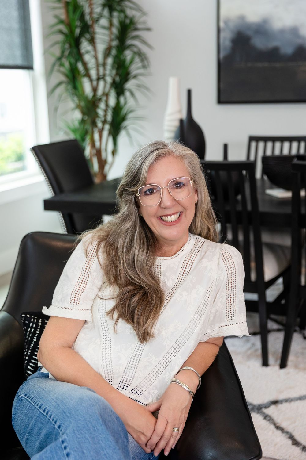 Smiling middle-aged woman with gray hair and glasses sitting in a modern black chair in a stylish living room.