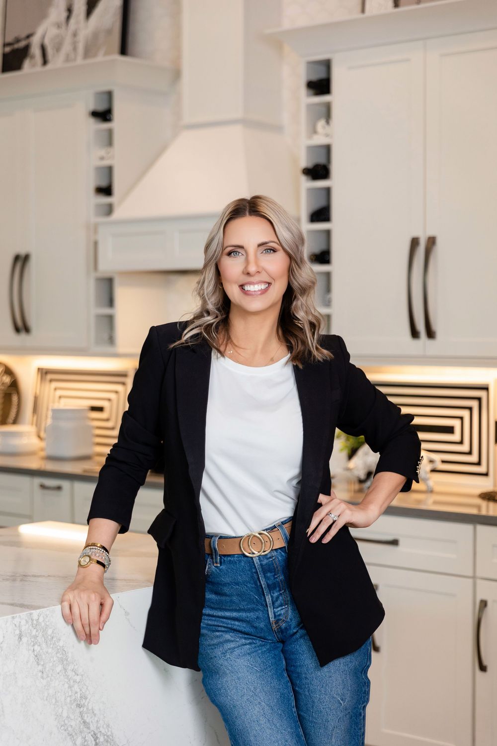 Woman in black blazer and jeans smiling in a modern white kitchen with marble countertops.