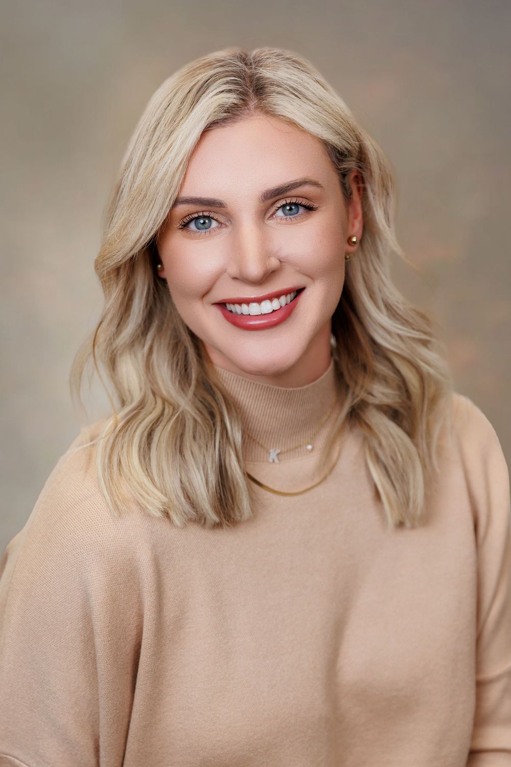 Smiling blonde woman with blue eyes wearing a beige turtleneck sweater and delicate necklace against neutral background.