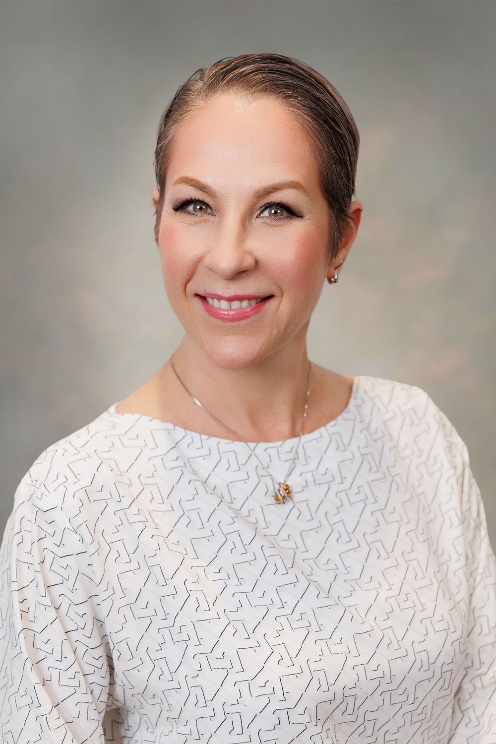 Professional headshot of a smiling woman with sleek hair, wearing a white patterned blouse and delicate gold necklace.