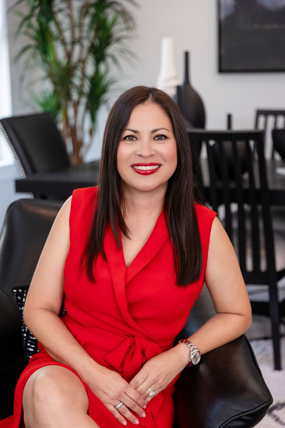 A smiling woman in a red sleeveless dress sits in a black chair in a modern interior setting.