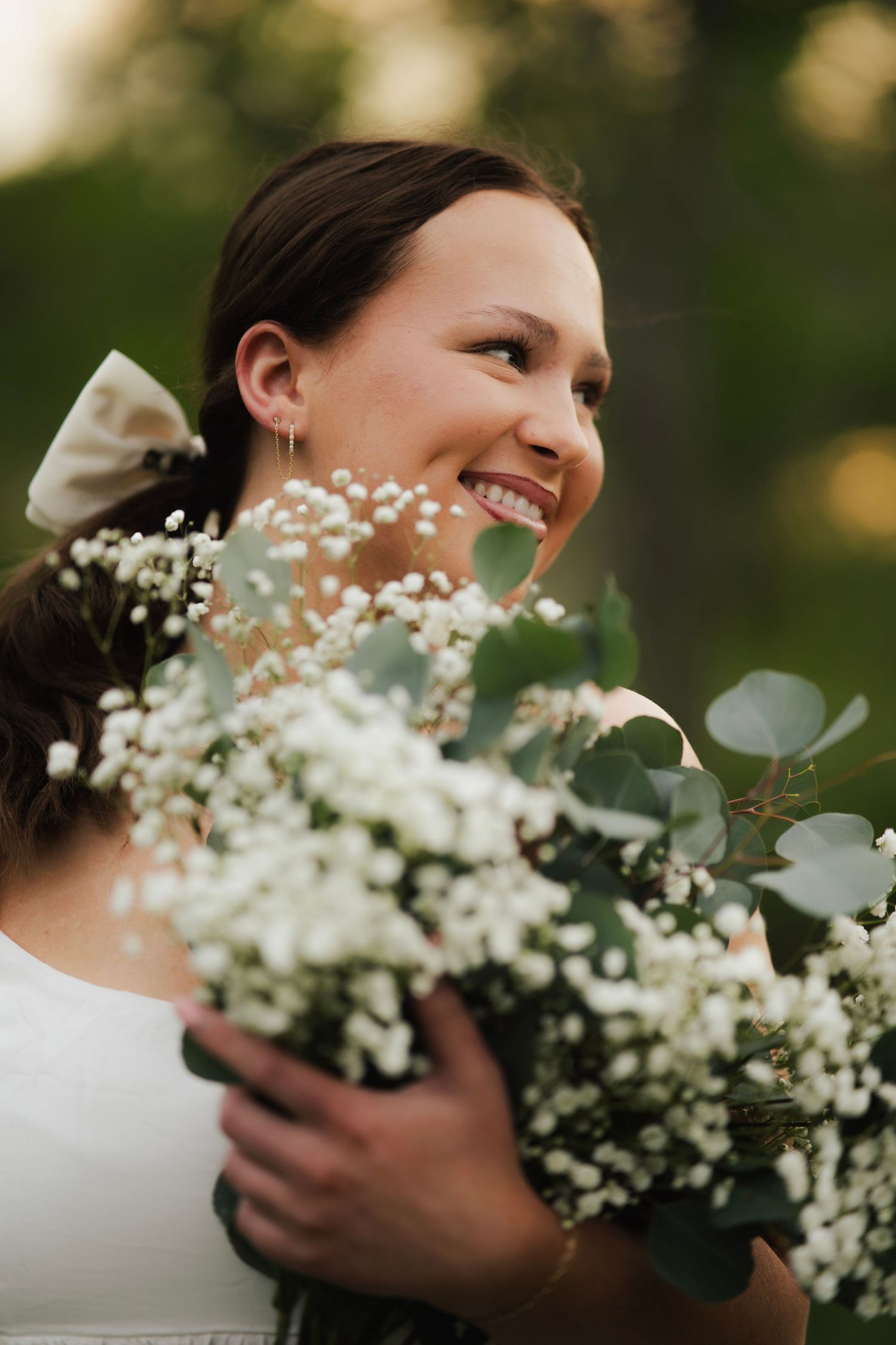 Smiling young woman holds a bouquet of white baby's breath and eucalyptus in a lush outdoor setting.