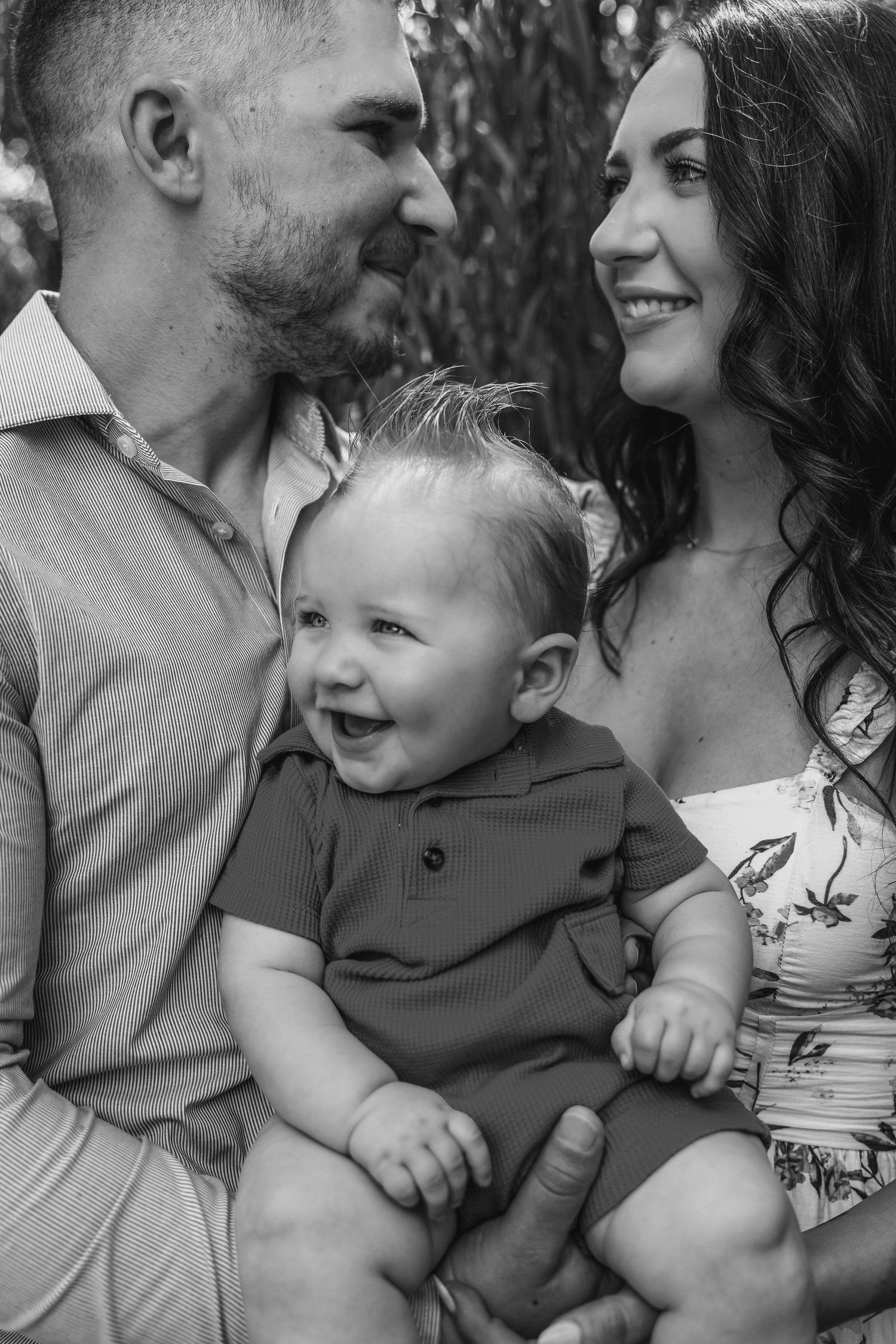 Black and white photo of smiling parents holding their laughing baby boy outdoors among trees.