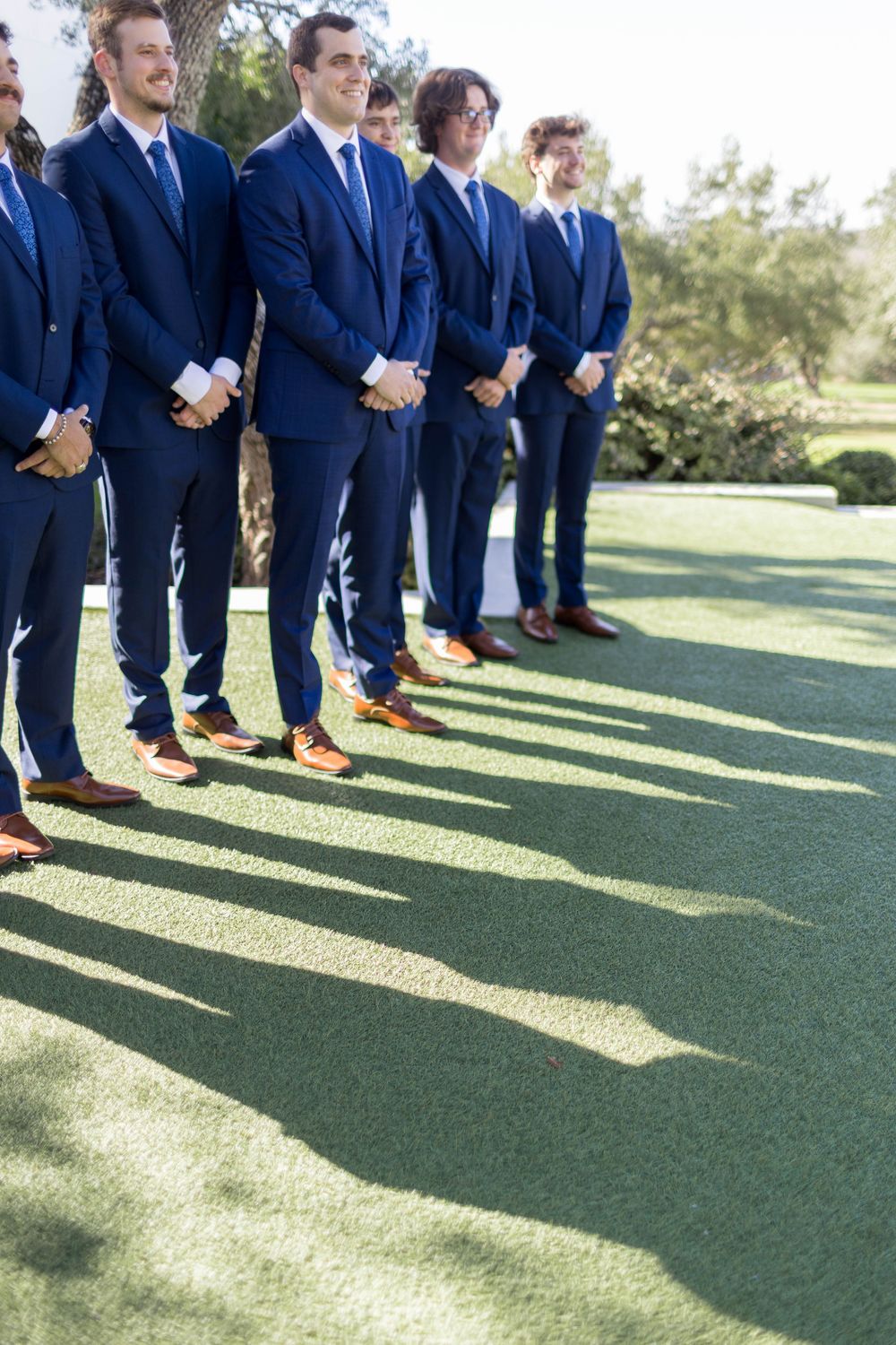 Groomsmen in navy blue suits and brown shoes standing in a row on green grass at an outdoor wedding ceremony.